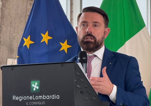 L'assessore Paolo Franco mentre parla con un microfono ad un convegno. In secondo piano le bandiere dell'UE e dell'Italia