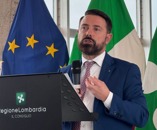 La foto ritrae l'assessore Paolo Franco mentre parla ad una conferenza, nello sfondo la bandiera dell'Eu, dell'Italia e della Lombardia