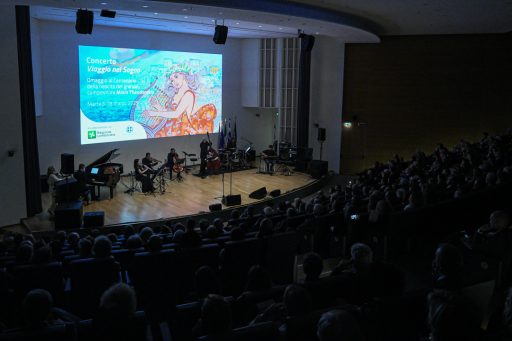 Il Teatro Testori preso dall'alto della scalinata, il pubblico è al buio e le luci illuminano il palco e i musicisti. Sullo sfondo la locandina dell'evento