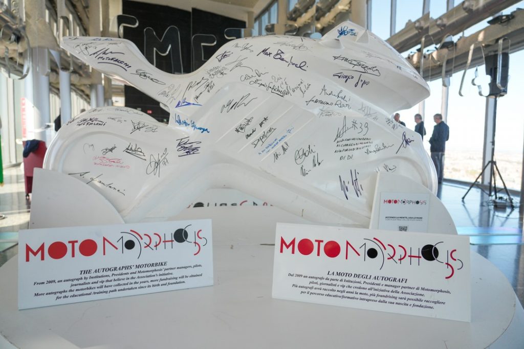 Una scultura di una moto di colore bianco, con diverse firme su tutto il mezzo. Alla base della moto il logo di Motomorphosis. Nello sfondo, lo specchio del 39esimo piano di Palazzo Lombardia