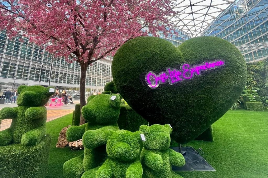 La foto ritrae una delle installazioni di Oasi Life Experience. Sono presenti degli orsacchiotti e un grande cuore ricoperti da un prato curato. Sul cuore c'è scritto con i led 'Oasi Life Experience'. Poi, è presente un albero di ciliegio fiorito
