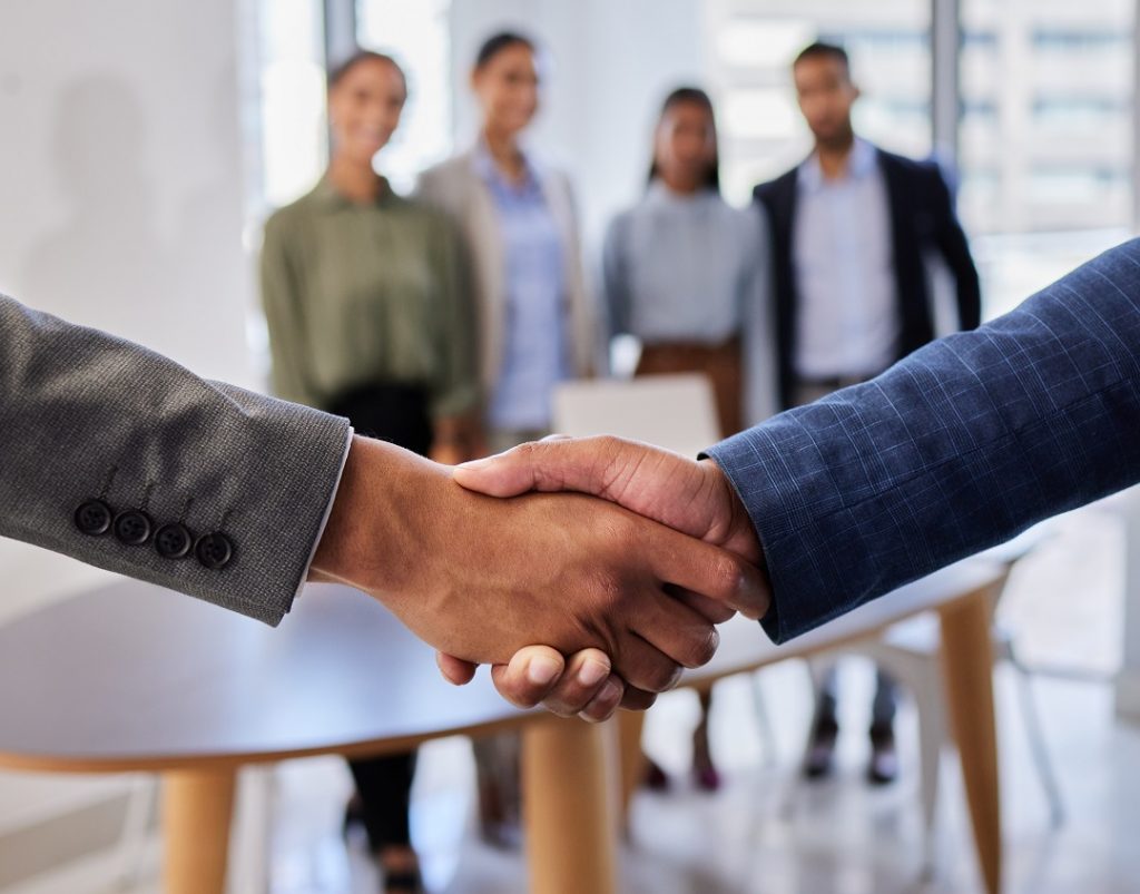 Stretta di mano in primo piano, dietro quattro persone sfumate. Foto utilizzata per l'articolo delle Case Aler Lodi con presidi territoriali e community manager