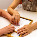 La foto ritrae le mani di una maestra che guidano le manine di una bambina, probabilmente ipovedente, su una braille board. Utilizzata per l'articolo dote scuola disabilità
