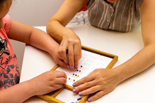 La foto ritrae le mani di una maestra che guidano le manine di una bambina, probabilmente ipovedente, su una braille board. Utilizzata per l'articolo dote scuola disabilità