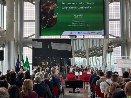 Locandina evento al Belvedere 'Per una rete delle miniere turistiche in Lombardia' proiettata. Panoramica della sala da dietro durante un intervento di Clerici