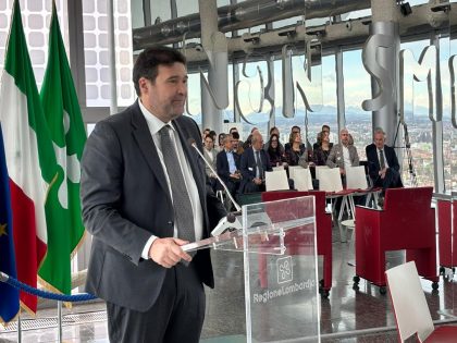 Assessore Giorgio Maione durante un suo intervento. Alle sue spalle le bandiere dell'UE, dell'Italia e della Lombardia. Foto utilizzata per articolo della pubblicazione della graduatoria bando SEED PA Lombardia