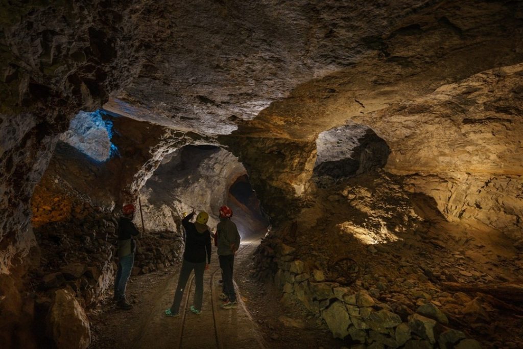 L'interno di una miniera con tre operatori. L'ambiente è buio e illuminato solo in alcuni punti, i tre operatori sono di spalle e si notano solo gli elmetti da lavoro