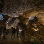 L'interno di una miniera con tre operatori. L'ambiente è buio e illuminato solo in alcuni punti, i tre operatori sono di spalle e si notano solo gli elmetti da lavoro