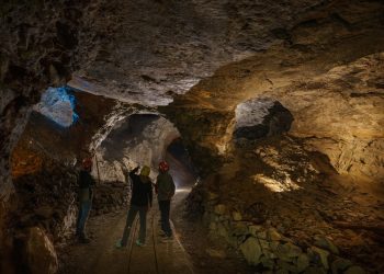 L'interno di una miniera con tre operatori. L'ambiente è buio e illuminato solo in alcuni punti, i tre operatori sono di spalle e si notano solo gli elmetti da lavoro