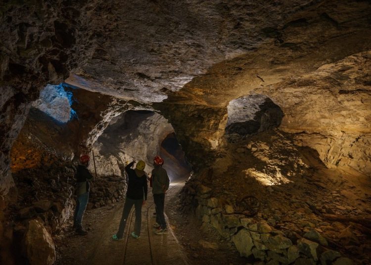 L'interno di una miniera con tre operatori. L'ambiente è buio e illuminato solo in alcuni punti, i tre operatori sono di spalle e si notano solo gli elmetti da lavoro