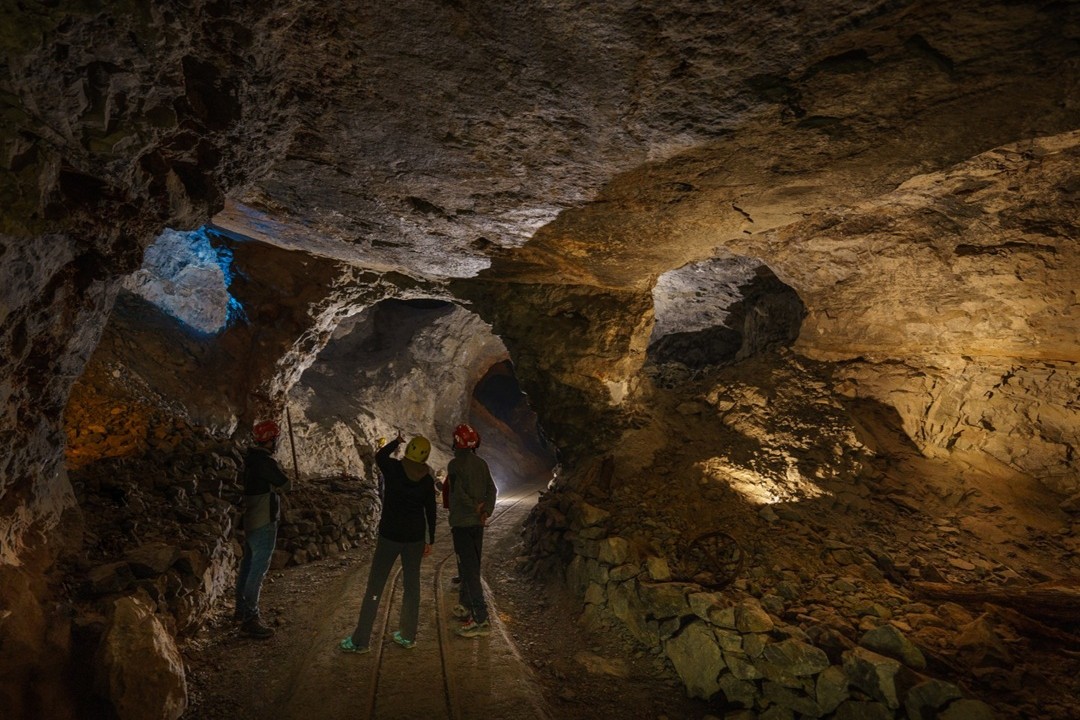 L'interno di una miniera con tre operatori. L'ambiente è buio e illuminato solo in alcuni punti, i tre operatori sono di spalle e si notano solo gli elmetti da lavoro