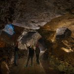 L'interno di una miniera con due operatori. L'ambiente è buio e illuminato solo in alcuni punti, i due operatori sono di spalle e si notano solo gli elmetti da lavoro