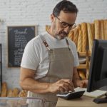 Commerciante che fa calcoli con calcolatrice davanti al pc. E' all'interno di una panetteria, alle sue spalle una lavagna nera con delle scritte bianche e accanto dele baguette e dei dolci. Foto utilizzata per i fondi di Regione Lombardia a sostegno dei Distretti del commercio