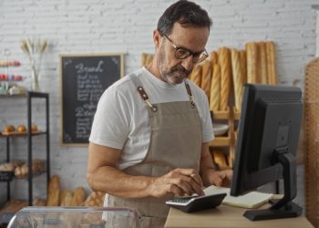Commerciante che fa calcoli con calcolatrice davanti al pc. E' all'interno di una panetteria, alle sue spalle una lavagna nera con delle scritte bianche e accanto dele baguette e dei dolci. Foto utilizzata per i fondi di Regione Lombardia a sostegno dei Distretti del commercio