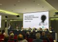 Convegno Associazione Ippocrate Salute mentale; l'immagine ritrae l'assessore La Russa durante il suo intervento; la visuale permette di vedere la locandina dell'evento trasmessa nel led e gli altri interlocutori seduti al tavolo.